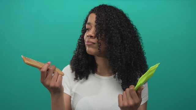 African american woman comparing two combs and checking curly hair texture in studio; choice decision uncertainty.