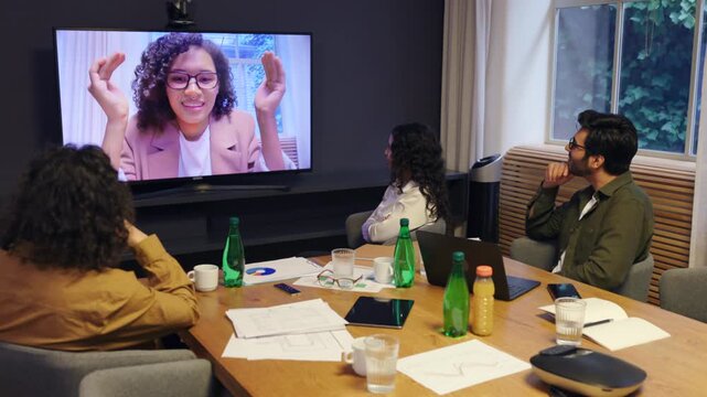Diverse coworkers watching female colleague on screen during online video conference in modern office showing teamwork presentation communication and professional remote collaboration
