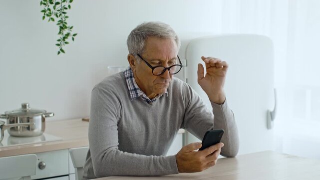 A senior man in his 60s sits at a kitchen table, focused on his smartphone. He wears glasses and is engaged in a casual activity at home, exuding a relaxed lifestyle.