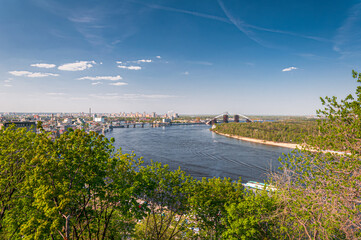 Panoramic view of Kyiv city skyline with bridges over the Dnipro River on a bright spring day. Clear blue sky, green trees, and calm water create a scenic urban landscape.
