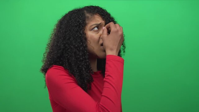 African american woman in red shirt with curly hair pinches nose and closes eyes in studio; disgust reaction.
