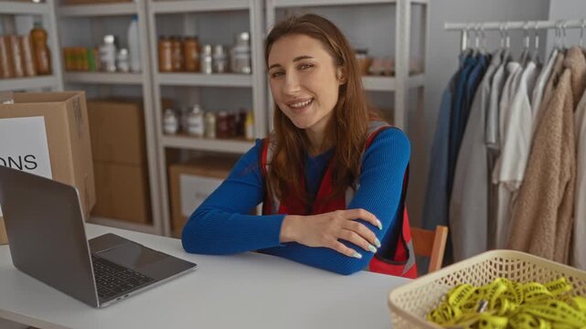 Young woman volunteer at a charity donation center sits near a laptop, surrounded by clothes and supplies, radiating warmth and dedication.
