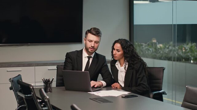 Two millennials discuss important business strategies while working on a laptop in a sleek conference room. The atmosphere is focused and industrious, reflecting a corporate lifestyle.