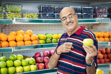 Fototapeta premium Senior indian man showing green apple fruit at fruit shop. Healthy lifestyle concept.