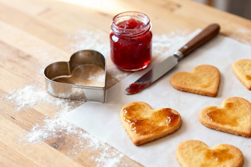 Freshly baked heart-shaped cookies are topped with sweet red jam, resting on baking paper beside a jar of jam, a knife, and a cookie cutter dusted with flour on a wooden board.