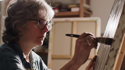 A focused senior woman with glasses diligently paints on a canvas with a brush in her art studio illuminated by natural light showcasing her passion for creative expression and artistic pursuit.