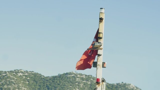 Close-up shot of the vibrant red Turkish national flag (Ay Yıldız) gently swaying on a flagpole against a backdrop of distant, lush green mountains and a clear blue sky.