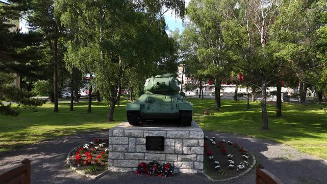 Aerial view of a T34 tank monument in a quiet park in Slovakia, surrounded by trees, grass and pathways. The preserved historic military vehicle stands on a stone pedestal in soft daylight.