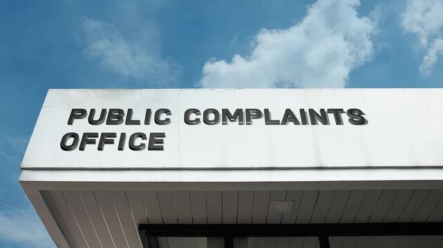 A Public Complaints Office word sign displayed on a civic or government building under a clear blue sky, symbolizing accountability, dispute resolution, citizen service, and official authority