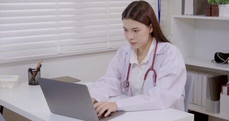 Asian female doctor wearing white coat and stethoscope working on laptop in clinic office, focused professional performing medical documentation, patient record management, online healthcare update - Powered by Adobe