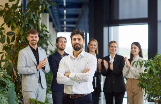 Fototapeta Confident business leader or ceo standing with arms crossed in modern office hallway while smiling professional colleagues applauding in background, celebrating success, achievements and teamwork.