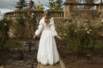 portrait of beautiful young blonde model wearing historical white wedding gown. ethereal back view figure, walking away in romantic rose garden of fairytale castle garden background © faestock