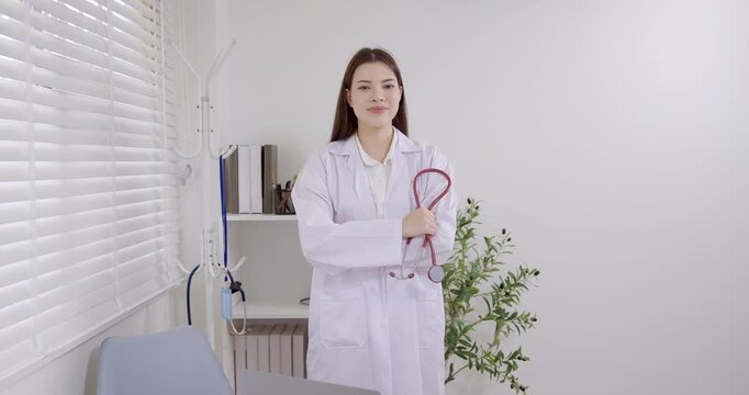 Asian female pediatrician standing in clinic office holding stethoscope with confidence, gentle smile, professional medical attitude, pediatric healthcare consultation child treatment slowmotion