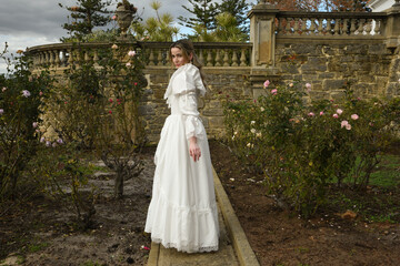 Fototapeta premium portrait of beautiful young blonde model wearing historical white wedding gown. ethereal back view figure, walking away in romantic rose garden of fairytale castle garden background