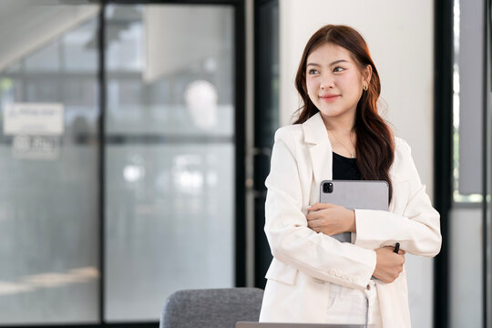 An elegant Asian businesswoman holding a digital tablet and looking forward with confidence in a modern office environment.