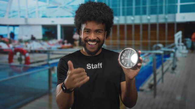 Man in staff uniform on cruise ship deck smiles holding clock and gesturing towards camera, set against a seaside outdoor background with pool and leisure area.