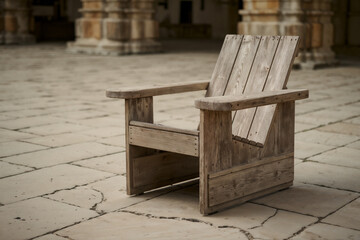 A weathered, rustic wooden Adirondack chair sits empty on a large, pale stone patio or courtyard,...