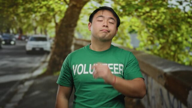 Young man wearing green volunteer shirt standing on urban street, looking thoughtful with hand on face, surrounded by trees and cars in the background.