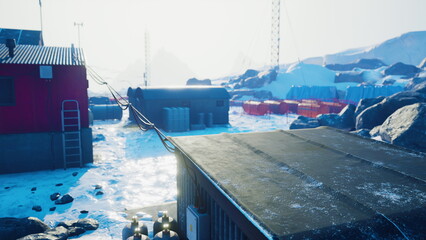 A vast icy expanse embraces a research station, where buildings sit among rocky terrain. Bright light reflects off the snow, illuminating the quiet, cold environment and equipment nearby.