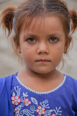 Serious little girl with brown hair in blue embroidered dress standing outdoors looking at the camera