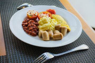 Hotel healthy breakfast menu of mashed potatoes, omelette, read beans, tomatoes, and sausages served on a white plate