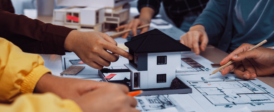 This image captures a collaborative design session where architects engage with a miniature house model, surrounded by blueprints and sketches, showcasing teamwork in architecture. SACTR
