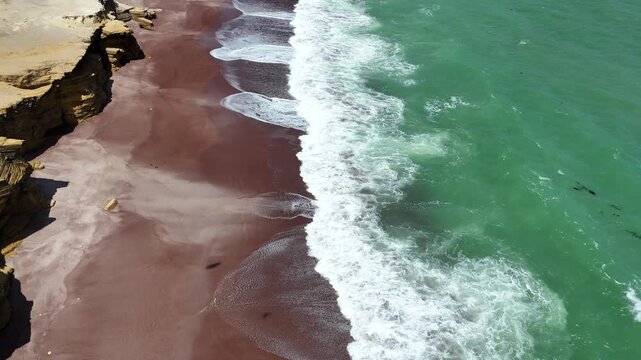 Close-up, face-down aerial shot captures the details of powerful white waves crashing onto the unique red sand beach of Playa Roja in the Paracas National Reserve