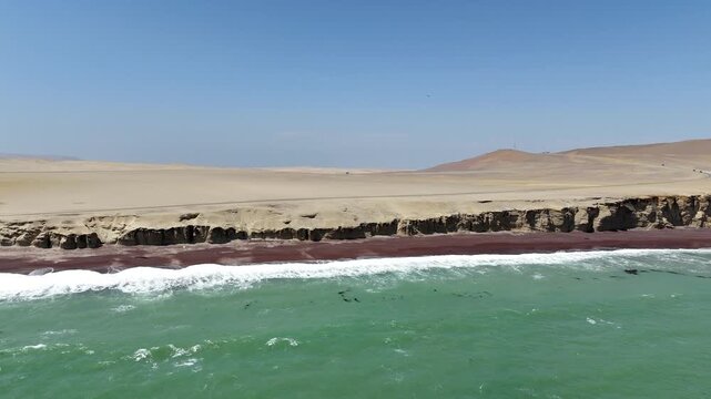 Pulling back from the iconic red sand Playa Roja beach in the Paracas National Reserve, Peru, showcasing heavy waves crashing against the dramatic desert coastline, cliffs, and turquoise ocean