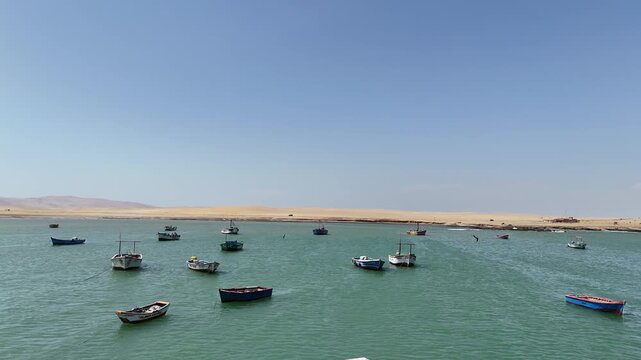 Moving straight towards a group of colorful traditional fishing boats anchored in the calm turquoise waters of Paracas Bay, Peru, with the arid desert coastline in the background