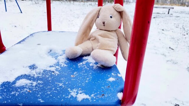 Lonely rabbit doll sitting alone on carousel at winter. Sad forgotten abandoned rabbit sitting outside in snowy day, lost toy waiting for owner. Loneliness concept, International missing Children day