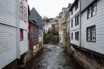 A street view of the historic town of Monschau, Germany, showcasing traditional half-timbered houses, including a prominent red-painted building, lining the banks of the Roer River.