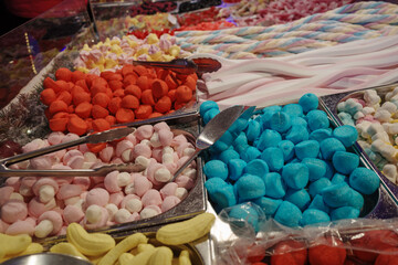 A close-up high-angle view of a pick and mix candy stall, showing metal bins filled with a variety of colorful sweets, including blue, red, and pink marshmallows, mushroom-shaped candies.