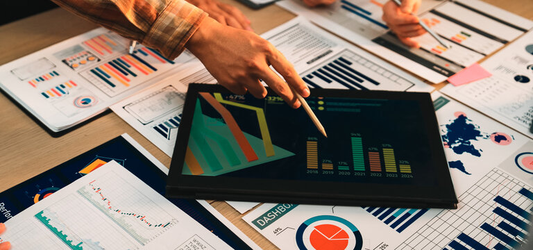 A diverse business team engages in collaborative data analysis using charts and graphs displayed on a tablet, surrounded by reports in a modern office setting. SACTR