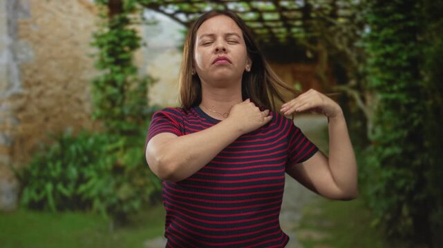 Woman clutching chest suggesting heartattack and holding neck at building entrance wearing red and navy striped shirt, pained expression, garden path visible; discomfort.