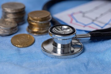 Stethoscope resting on a blue cloth with stacks of coins and a financial graph, symbolizing healthcare costs and medical finances