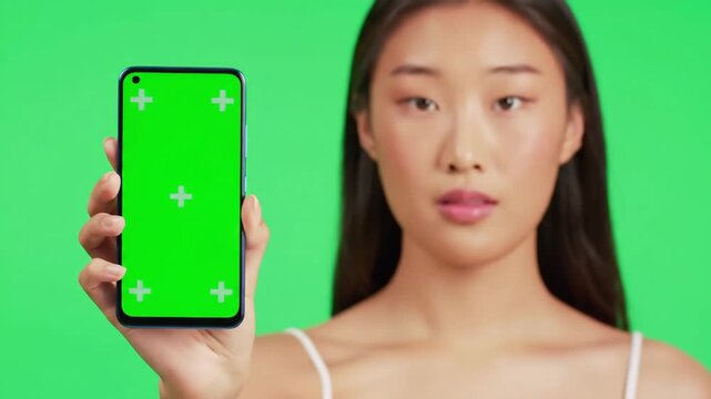 Young Asian Woman Holding Smartphone with Green Screen Displaying Tracking Markers for Easy Replacement Standing Against a Bright Green Background.