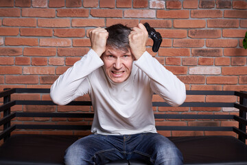 A middle-aged man gamer holds a gamepad and plays an online game on a computer.