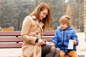 Mother and Son Folding Paper Boat Together on Park Bench During Autumn Day