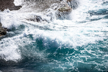 Clear turquoise sea water near rocky coastline. Sea with stones underwater. Coastal landscape with blue wave. Tropical seashore with ocean water. Rocky coast. Blue sea surface with foam waves.
