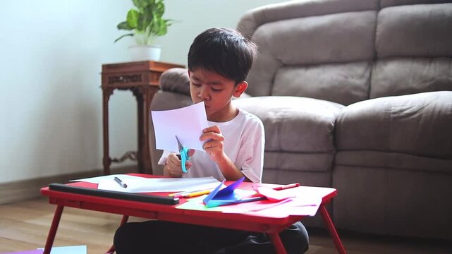 Focused Young asian boy on creative paper craft project while cutting with scissors at home
