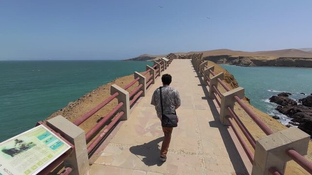 DIY Destinations TV host Charles Huang walks along the paved path and railing of the observation viewpoint in the Paracas National Reserve, Peru, overlooking the blue Pacific Ocean