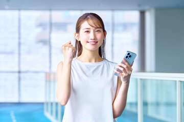 ジムでトレーニングしてスマホを見て喜ぶ女性　撮影協力：スポーツクラブ&サウナスパ ルネサンス 幕張24