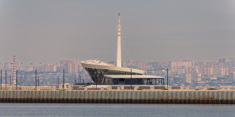 Ruby 360 Restaurant, a modern architectural landmark, stands by a pier with Baku, Azerbaijan's city skyline in the hazy distance.
