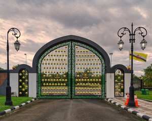 Side entrance of Jame Asr Hassanil Bolkiah Mosque, Bandar Seri Begawan, Brunei Darussalam. Ornate green and gold gate with street lamps and Brunei flag under cloudy sky.