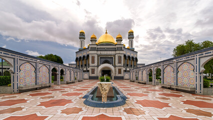 Jame' Asr Hassanil Bolkiah Mosque, with golden domes, minarets, and ornate mosaic walls, a fountain, and patterned courtyard under a cloudy sky in Bandar Seri Begawan, Brunei.