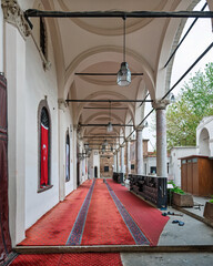 Exterior view of Hisar Mosque in Izmir, Turkey. Arched corridor with red prayer carpets, classical columns, and hanging lanterns. A Turkish flag is displayed in a window