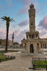 Izmir Clock Tower and Yali Mosque in Konak Square, Izmir, Turkey, with a prominent palm tree and pigeons, under a colorful cloudy sky at dusk