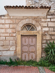 Historic ornate metal door with arched window, crescent moon carving, and tiled roof on a stone wall. Green plants, brick pavement