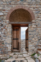 Historic stone and brick wall with an arched entrance. An old wooden door, ajar, reveals a rugged, rocky path under a cloudy sky. Rustic architectural detail