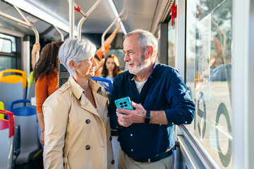 Senior couple standing and talking on a public bus, enjoying transit mobility connection. Man holding a mobile phone